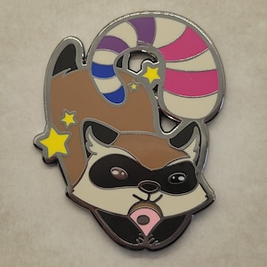 May include: A silver enamel pin featuring a cartoon raccoon wearing a candy cane hat and holding a pink donut. The raccoon has brown fur, black eyes, and a white muzzle. The candy cane is purple, blue, and white. The pin has a shiny finish.