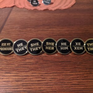 May include: Seven black and gold round pins with different pronoun options. The pins read "Ask My Pronouns", "He/They", "She/They", "She/Her", "He/Him", "Xe/Xem", and "They/Them".