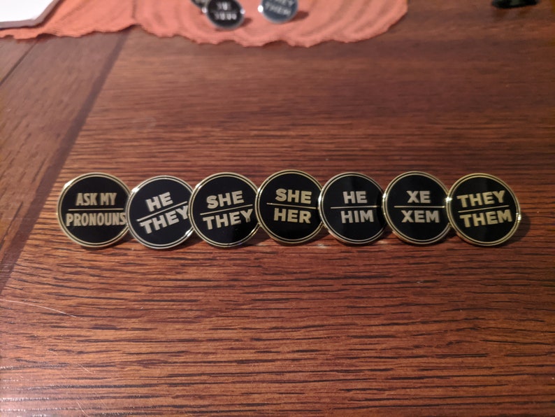 May include: Seven black and gold round pins with different pronoun options. The pins read "Ask My Pronouns", "He/They", "She/They", "She/Her", "He/Him", "Xe/Xem", and "They/Them".