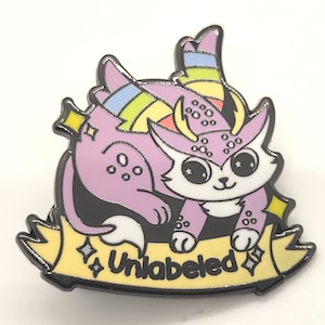 May include: A purple and white enamel pin featuring a cartoon creature with rainbow horns and a yellow banner that reads "Unlabeled".