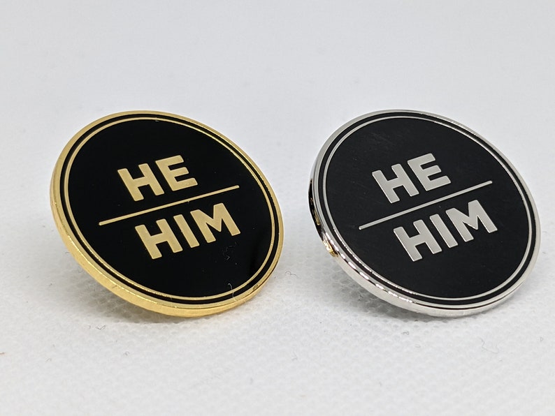 May include: Two round pins with black backgrounds and gold lettering. The text on the pins reads "HE HIM". One pin has a gold border and the other has a silver border.