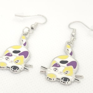 May include: A pair of silver dangle earrings featuring a cartoon cat design. The cat is white with yellow, black, and purple spots. The cat is looking to the left.