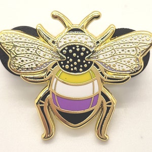 May include: Gold-colored bee pin with black and white wings and a yellow, white, and purple striped abdomen. The bee is a symbol of the non-binary community.