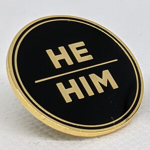 May include: A gold-colored circular pin with a black background and the words "HE/HIM" in gold lettering.