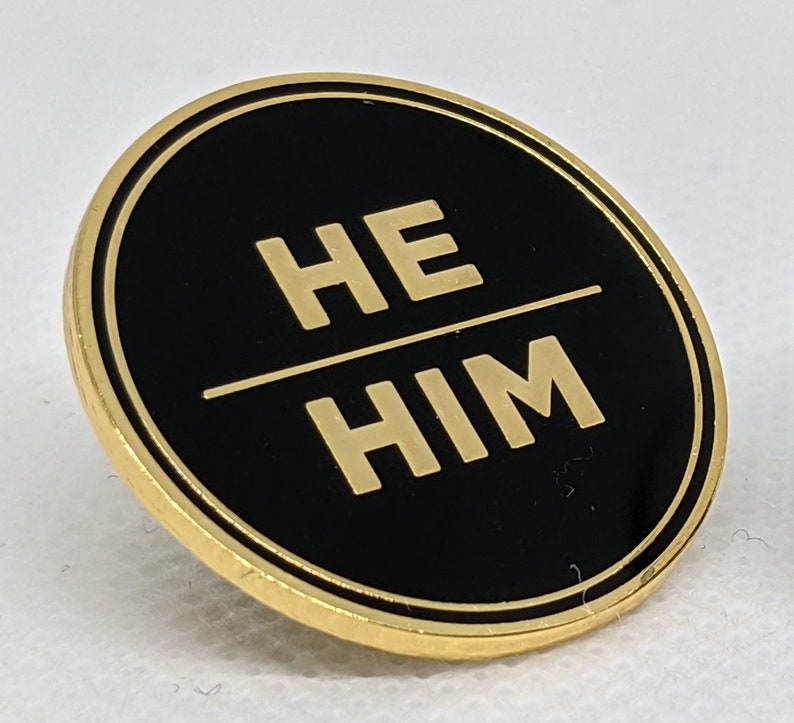 May include: A gold-colored circular pin with a black background and the words "HE/HIM" in gold lettering.
