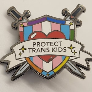 Może przedstawiać: Srebrna szpilka emaliowana z wzorem tarczy. Tarcza jest ozdobiona tęczowym sercem i napisem "PROTECT TRANS KIDS" na białym banerze. Dwa miecze są skrzyżowane za tarczą.