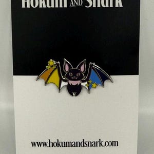 May include: A metal enamel pin featuring a black bat with pink accents and wings colored in yellow, orange, blue, and dark blue, decorated with small yellow stars. The pin is attached to a black and white display card labeled Hokum and Snark.