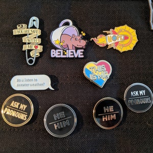 May include: A collection of enamel pins featuring various designs, including a safety pin with the text "You are safe with me", a hippopotamus in a teacup with the text "Believe", a heart with the text "This is me", a rocket with the text "Nope", and several pins with the text "Ask my pronouns" and "He Him".