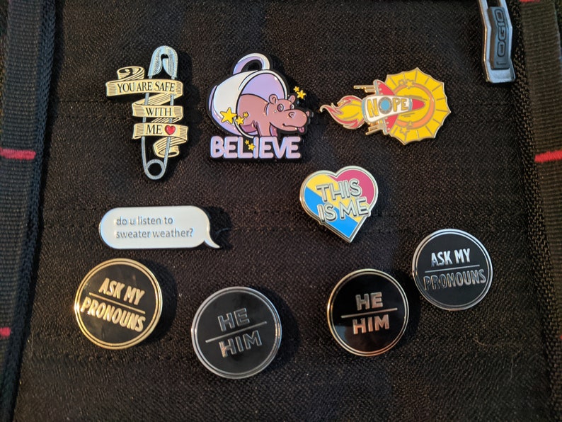 May include: A collection of enamel pins featuring various designs, including a safety pin with the text "You are safe with me", a hippopotamus in a teacup with the text "Believe", a heart with the text "This is me", a rocket with the text "Nope", and several pins with the text "Ask my pronouns" and "He Him".
