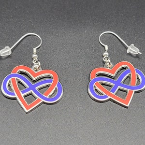 Op de afbeelding: Een paar zilveren oorbellen met een rood en blauw oneindigheidssymbool dat is verweven met een hartvorm.