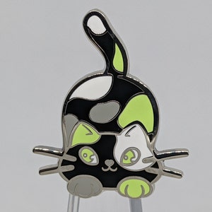 May include: A black, white, and green enamel pin of a cartoon cat with a spotted tail. The cat has a black nose, green eyes, and a white patch on its face.