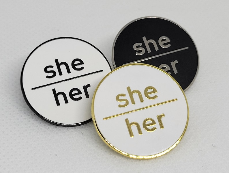 K&ouml;nnte beinhalten: Drei runde Pins mit wei&szlig;em Hintergrund und schwarzen R&auml;ndern. Die Pins zeigen "she/her" in schwarzer Schrift auf wei&szlig;em Hintergrund, "she/her" in goldener Schrift auf wei&szlig;em Hintergrund und "she/her" in wei&szlig;er Schrift auf schwarzem Hintergrund.