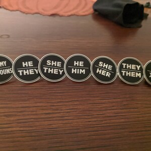 May include: A set of eight round black and white pins with different pronoun options. The pins read "Ask My Pronouns", "He/They", "She/They", "He/Him", "She/Her", "They/Them", and "Xe/Xem".
