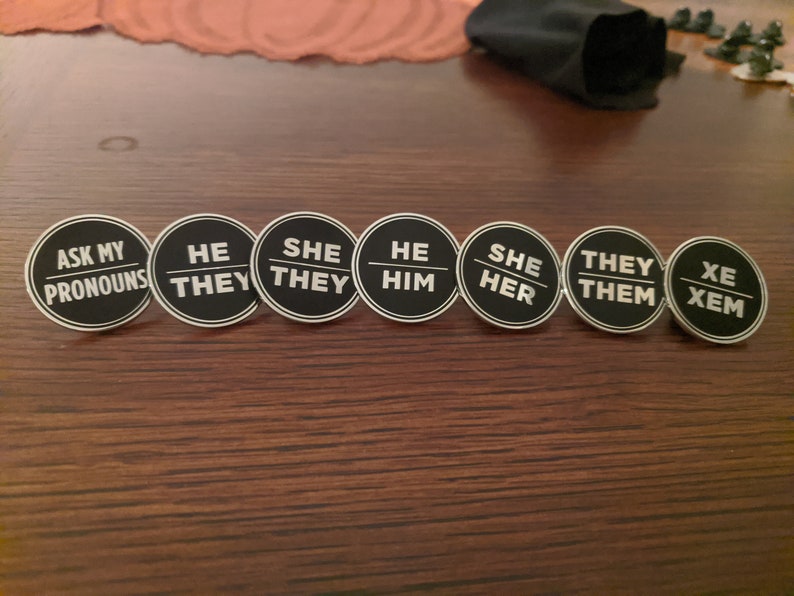 May include: A set of eight round black and white pins with different pronoun options. The pins read "Ask My Pronouns", "He/They", "She/They", "He/Him", "She/Her", "They/Them", and "Xe/Xem".