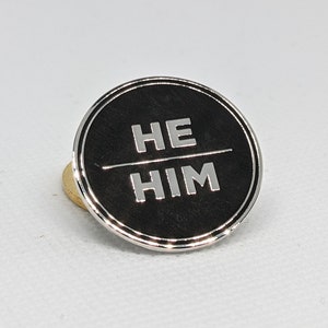 May include: A black circular pin with a silver border and the words "HE HIM" in white lettering.