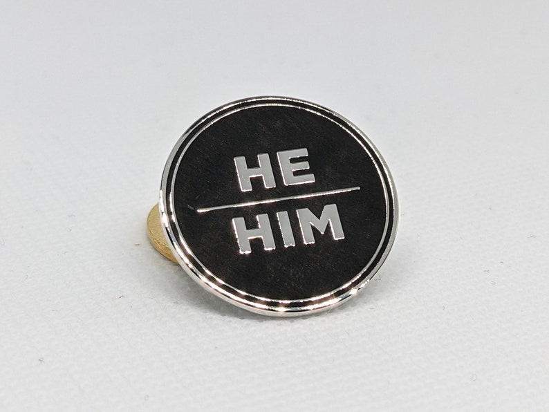 May include: A black circular pin with a silver border and the words "HE HIM" in white lettering.