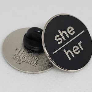 K&ouml;nnte beinhalten: Schwarzer Emaille-Pin mit silberner Beschriftung "she/her". Der Pin ist an einer silbernen R&uuml;ckseite mit einer Sicherheitsnadel befestigt.