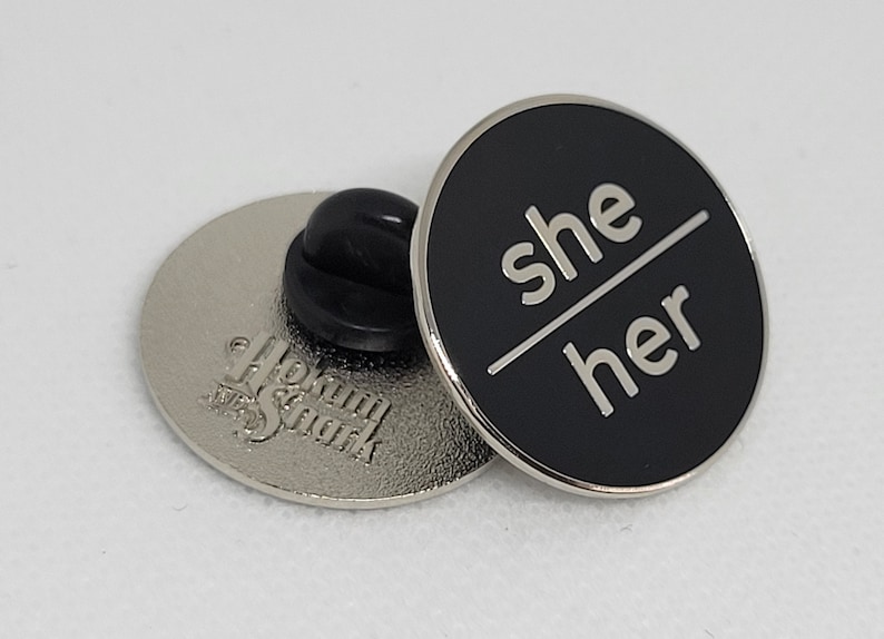 K&ouml;nnte beinhalten: Schwarzer Emaille-Pin mit silberner Beschriftung "she/her". Der Pin ist an einer silbernen R&uuml;ckseite mit einer Sicherheitsnadel befestigt.