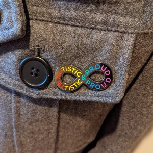 K&ouml;nnte beinhalten: Eine schwarze Unendlichkeits-Symbol-Anstecknadel mit den Worten "Autistisch & Stolz" in Regenbogenfarben darauf. Die Anstecknadel ist an einer grauen Jacke befestigt.