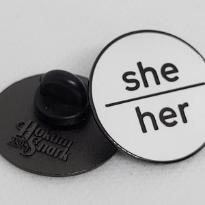 K&ouml;nnte beinhalten: Eine wei&szlig;e Emaille-Anstecknadel mit schwarzem Text, der "she/her" lautet. Die Nadel ist rund und hat eine silberne R&uuml;ckseite mit einer Sicherheitsnadel.