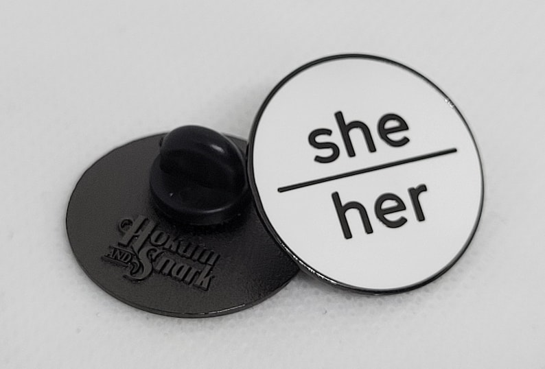 K&ouml;nnte beinhalten: Eine wei&szlig;e Emaille-Anstecknadel mit schwarzem Text, der "she/her" lautet. Die Nadel ist rund und hat eine silberne R&uuml;ckseite mit einer Sicherheitsnadel.