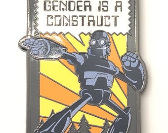 Op de afbeelding: Een zilveren metalen pin met een retro-stripboekontwerp. Een robot met een spraakballon die "GENDER IS A CONSTRUCT" zegt, staat voor een gele en oranje zonnestralenachtergrond met bomen aan de onderkant.