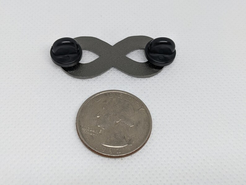 K&ouml;nnte beinhalten: Eine silberne Unendlichkeits-Symbol-Anstecknadel mit schwarzen Gummibeschl&auml;gen. Die Anstecknadel ist etwa so gro&szlig; wie eine US-amerikanische 25-Cent-M&uuml;nze.
