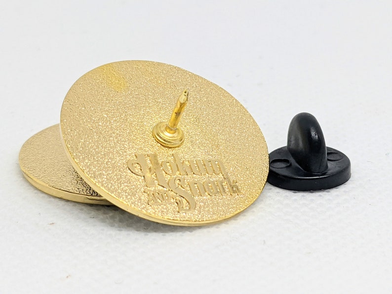 K&ouml;nnte beinhalten: Goldfarbener Pin mit schwarzer Gummur&uuml;ckseite. Der Pin tr&auml;gt den Text "Wakun" und "Snack" in einer stilisierten Schrift.