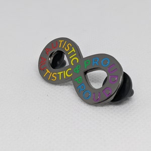 K&ouml;nnte beinhalten: Eine schwarze Metallstiftschleife in Form eines Unendlichkeitszeichens mit den Worten "Autistic & Proud" in Regenbogenfarben.