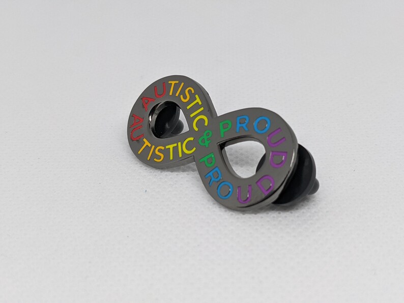 K&ouml;nnte beinhalten: Eine schwarze Metallstiftschleife in Form eines Unendlichkeitszeichens mit den Worten "Autistic & Proud" in Regenbogenfarben.