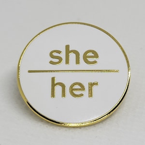 K&ouml;nnte beinhalten: Runder wei&szlig;er Pin mit goldener Umrandung. Der Pin tr&auml;gt die Aufschrift "she/her" in goldenen Buchstaben.
