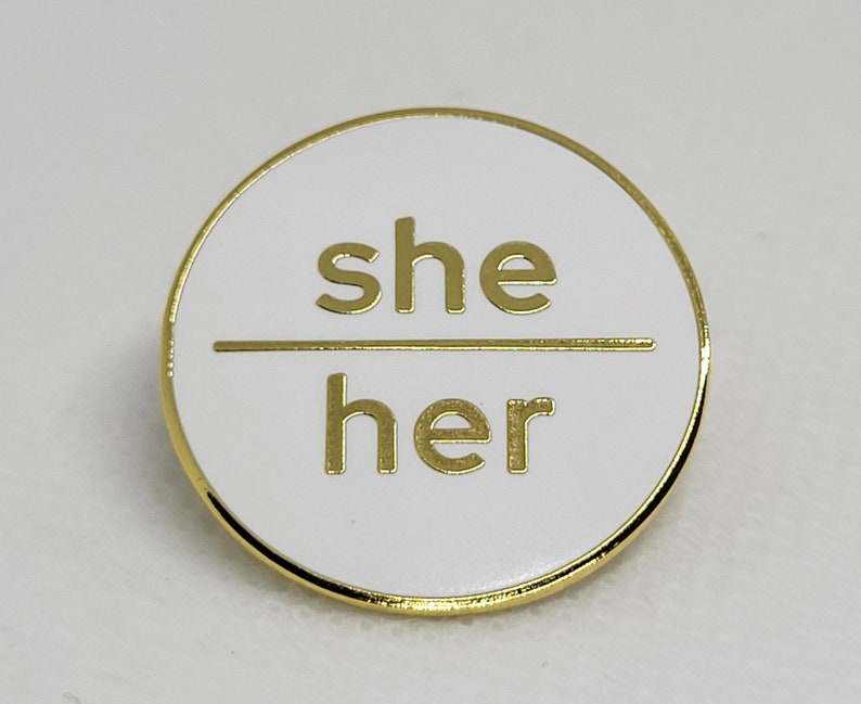 K&ouml;nnte beinhalten: Runder wei&szlig;er Pin mit goldener Umrandung. Der Pin tr&auml;gt die Aufschrift "she/her" in goldenen Buchstaben.