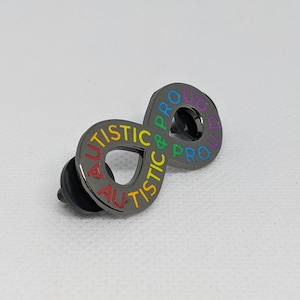 K&ouml;nnte beinhalten: Eine schwarze Metallstiftschleife in Form eines Unendlichkeitszeichens mit den Worten "Autistic & Proud" in Regenbogenfarben.