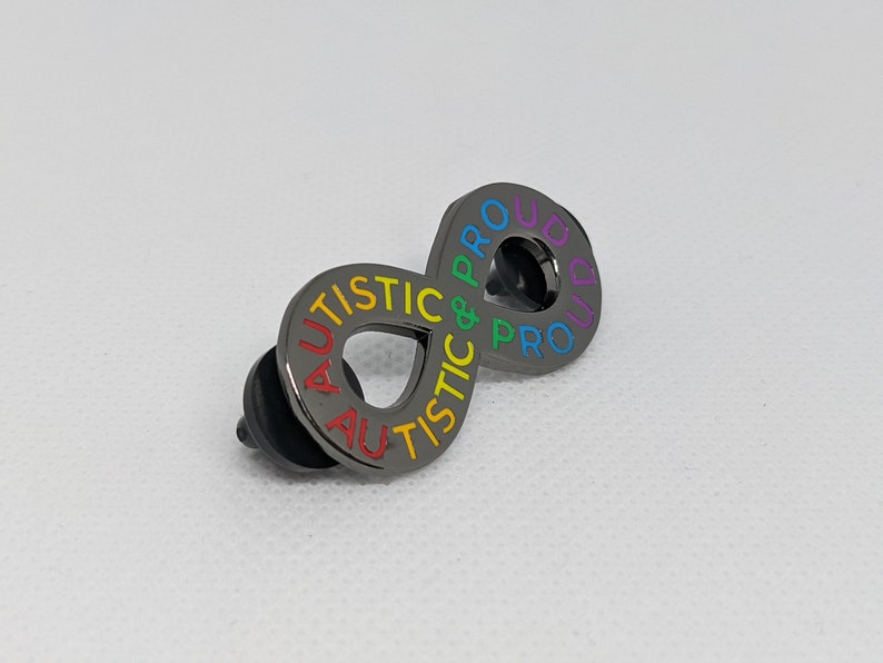 K&ouml;nnte beinhalten: Eine schwarze Metallstiftschleife in Form eines Unendlichkeitszeichens mit den Worten "Autistic & Proud" in Regenbogenfarben.