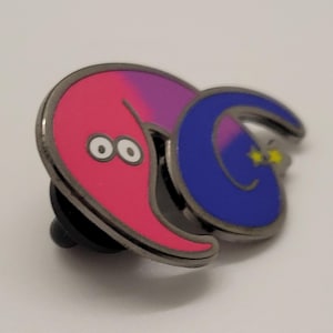 May include: A pink and purple worm with a blue worm wrapped around it. The blue worm has a yellow star on its tail. Both worms have black outlines and white eyes. The worms are attached to a metal pin.