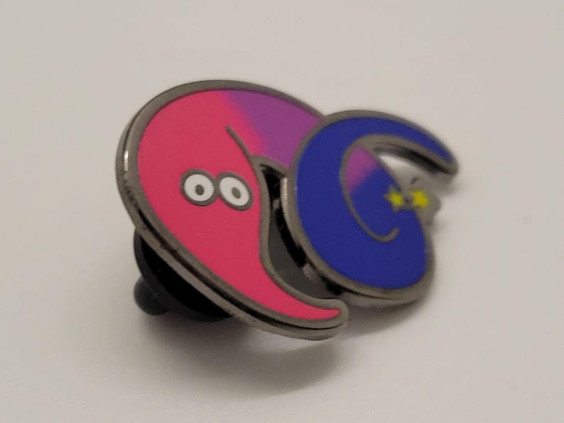 May include: A pink and purple worm with a blue worm wrapped around it. The blue worm has a yellow star on its tail. Both worms have black outlines and white eyes. The worms are attached to a metal pin.