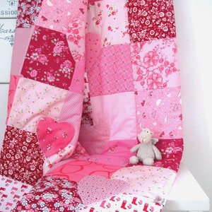 Könnte beinhalten: Eine rosafarbene und rote Patchwork-Decke mit einem herzförmigen Design. Die Decke ist gefaltet und über einen weißen Stuhl gelegt. Ein kleines Stofftier sitzt auf der Decke.