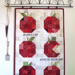 Könnte beinhalten: Ein weißer Wandbehang aus Stoff mit einem roten, weißen und grünen Patchwork-Design aus Äpfeln. Der Text "An apple a day keeps the doctor away" ist auf den Stoff gestickt.