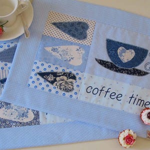 Könnte beinhalten: Blaue, gesteppte Tischsets mit Patchwork-Designs. Eines zeigt eine Kaffeetassen-Applikation mit einem Herz und den Worten "coffee time". Das andere hat blau-weiß gemusterte Patches. Eine weiße Tasse und Untertasse sind im Hintergrund.