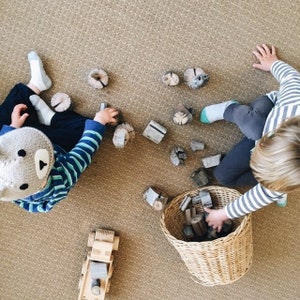 May include: Two children play with wooden blocks and a toy truck on a carpeted floor. One child wears a brown and white knitted hat. The other child is holding a wicker basket filled with more wooden blocks.