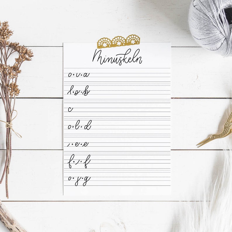 May include: A white sheet of paper with lined writing space and the word "Minuskteln" at the top. The paper is taped to a white wooden surface. The letters of the alphabet are written in cursive on the paper.