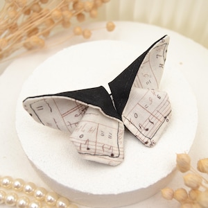 Könnte beinhalten: Eine schwarze und weiße, schmetterlingsförmige Haarklammer mit einem Notenmuster. Die Klammer ist aus Stoff und hat eine schwarze Rückseite.