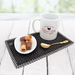 Könnte beinhalten: Eine weiße Keramiktasse mit einer schwarzen Katzensilhouette und dem Text "Kleines  Man" in Rosa. Die Tasse steht auf einem schwarzen und weißen gesteppten Untersetzer mit einer weißen Untertasse und Schokoladen-Waffel-Keksen.