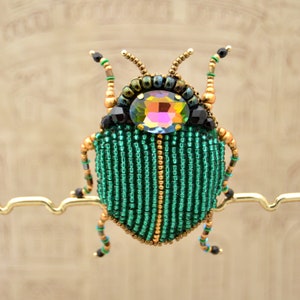 Könnte beinhalten: Eine grüne und goldene Perlen-Käfer-Brosche mit einem großen, schillernden Juwel in der Mitte. Die Brosche ist an einem goldenen Metallständer befestigt.