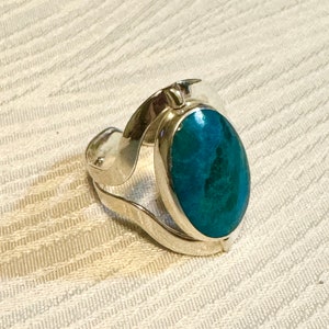 May include: A silver ring featuring an oval, teal-colored stone. The ring has a unique, flowing design with a band that curves around the finger. The stone's color varies from deep teal to lighter shades of blue and green.