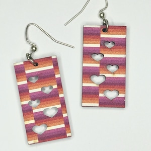 Op de afbeelding: Een paar rechthoekige oorbellen met een gestreept patroon in tinten rood, oranje en paars. Elke oorbel heeft vier hartvormige uitsparingen. De oorbellen hebben zilverkleurige oorhaakjes.