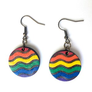 Op de afbeelding: Ronde oorbellen in regenboogkleuren met een golvend patroon en glitter. De oorbellen zijn gemaakt van hout en hebben bronzen haakjes.