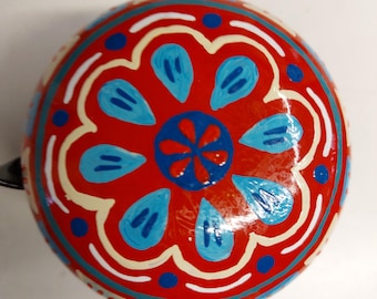 Peut inclure: Une sonnette de vélo ronde avec une base rouge vive ornée d'un motif floral peint à la main. Le design comprend un cercle bleu central avec des pétales rouges, entouré de formes en goutte d'eau bleu clair, de contours blancs et de points bleu foncé.