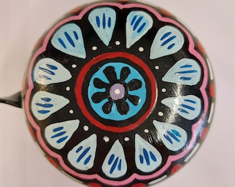Peut inclure: Une sonnette de vélo peinte à la main avec un motif floral. La cloche présente un fond noir avec un cercle bleu central et une fleur violette. Le motif comprend des formes de goutte d'eau bleu clair et une bordure rose et rouge.