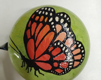 Peut inclure: Objet rond vert peint à la main, orné d'un motif de papillon monarque. Le papillon a des ailes orange et noires avec des points blancs. L'objet a une tige noire et est probablement un objet décoratif.